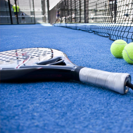 Close-up view of a padel racket and three yellow padel balls on a blue court, with a net in the background. The grip of the racket is prominent in the foreground.