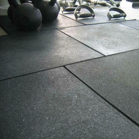 Close-up view of a gym floor with large black rubber mats. Kettlebells and other exercise equipment are visible in the background. The image focuses on the flooring texture and arrangement of the mats.