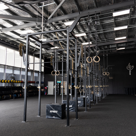 Interior of a gym with CrossFit equipment, including pull-up bars, gymnastic rings, and various weights along the wall. The space has high ceilings with metal beams and large windows, letting in natural light.