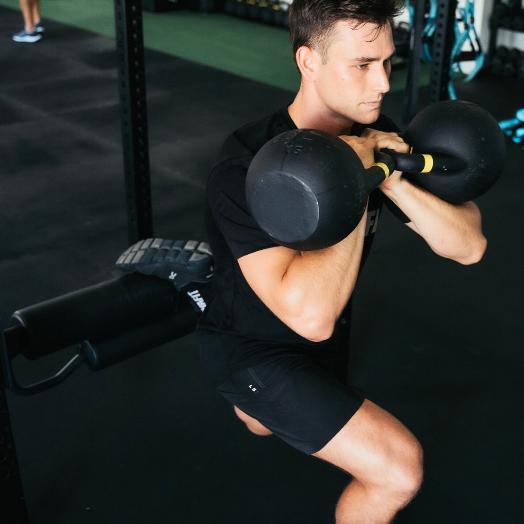 A man performs a weighted lunge in a gym, holding kettlebells at shoulder height. He wears a black shirt and black shorts, concentrating on his exercise. Gym equipment is visible in the background.