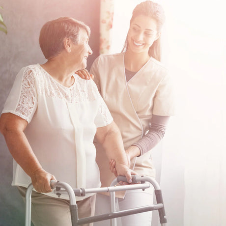 A caregiver assisting an elderly woman with a walker. The elderly woman is smiling and looking at the caregiver, who is also smiling. They appear to be in a well-lit room with sunlight streaming in.