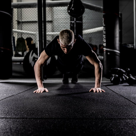 A person with short blond hair does push-ups on a black mat in a dimly lit gym. In the background, there is a boxing ring and gym equipment. The atmosphere is serious and focused.