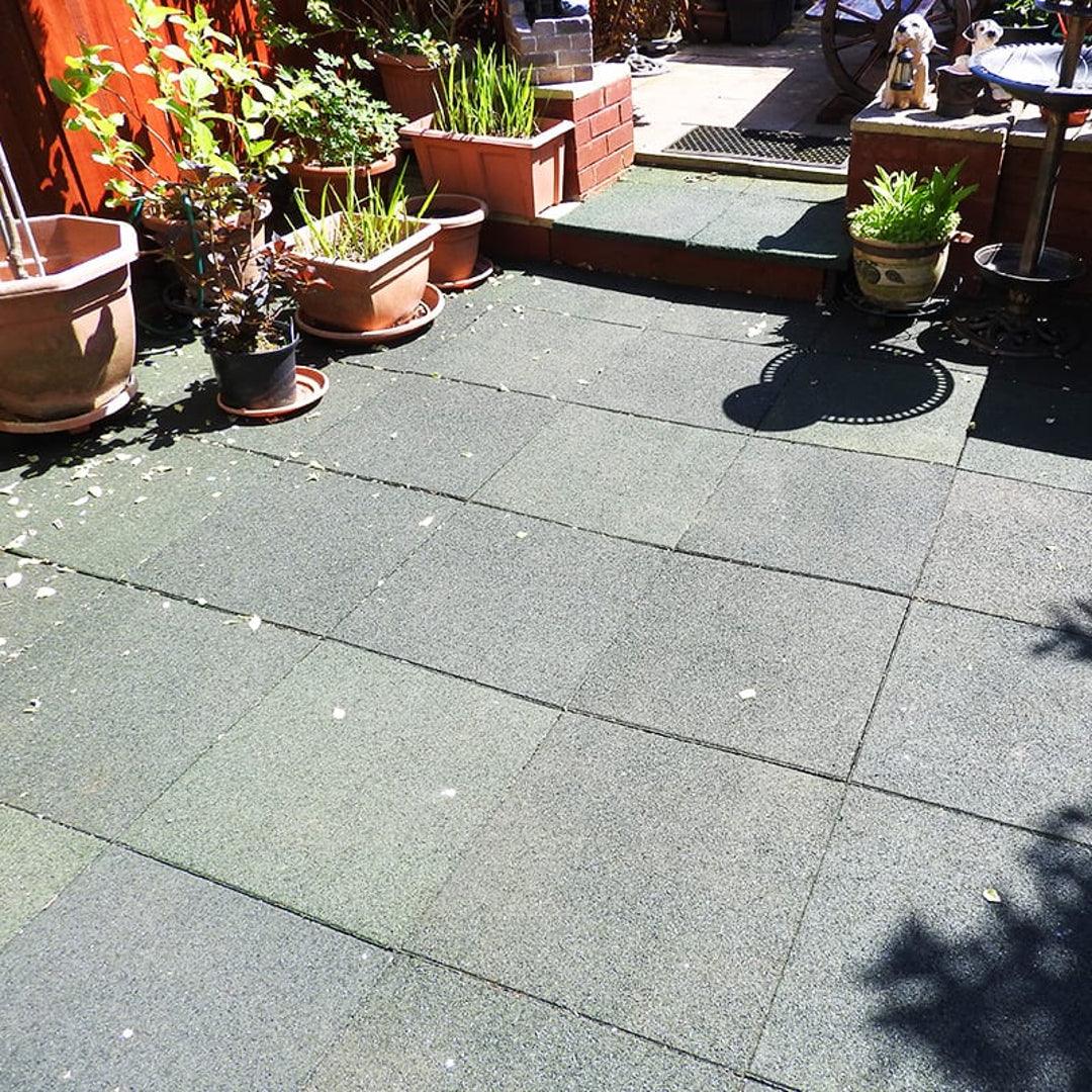 A sunlit garden patio with green tiles, various potted plants, and a small metal table with a cat statue. Steps lead to a raised area with more plants and another statue visible in the background.