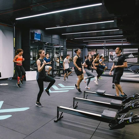 A group of people are exercising in a spacious gym. They're doing high knee exercises led by an instructor. Various gym equipment is visible, including rowing machines, weights, and a TV screen in the background.